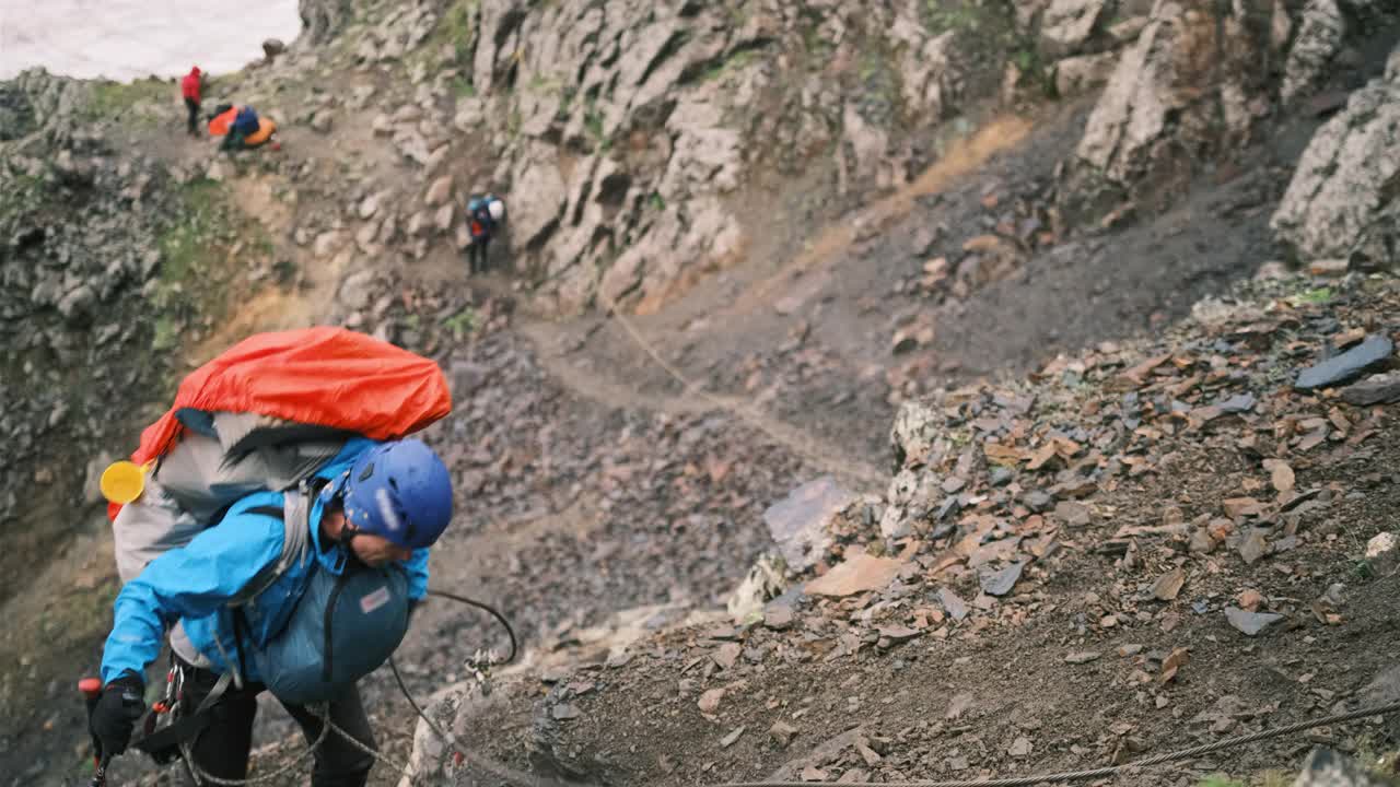 一位上了年纪的登山者背着背包爬上了山，背包里装着登山用的设备和断裂的杆子，用一个带安全网的锁扣固定在绳子上。视频素材