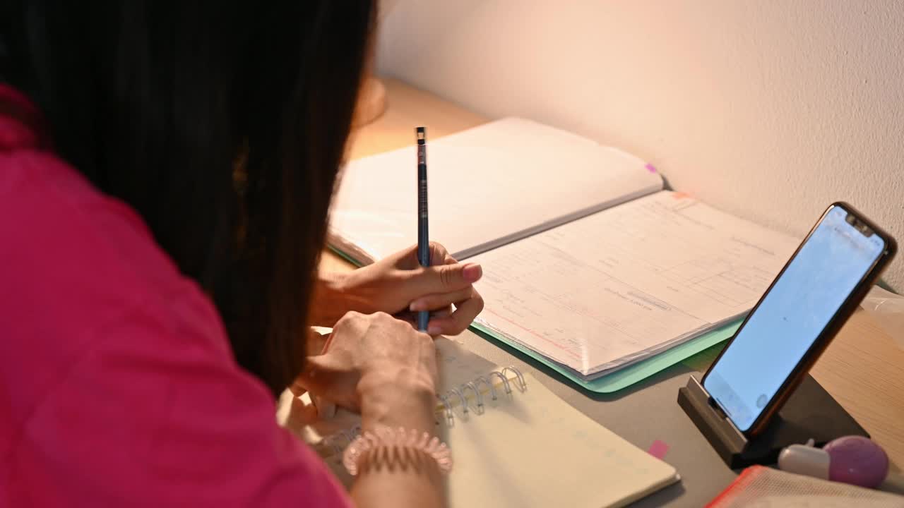 亚洲女性通过智能手机在线学习，并在家里的桌子上记笔记视频素材