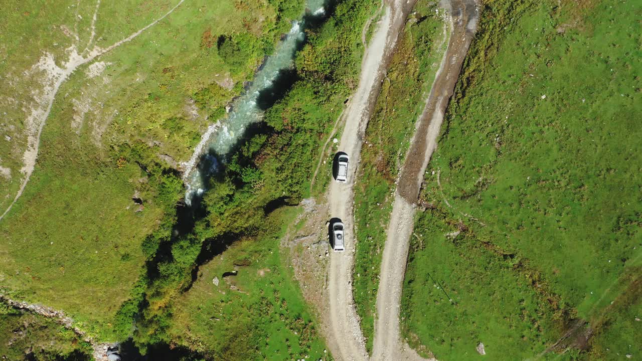 从上而下的鸟瞰图，两辆汽车沿着山间河流的土路驶向大桥。视频素材