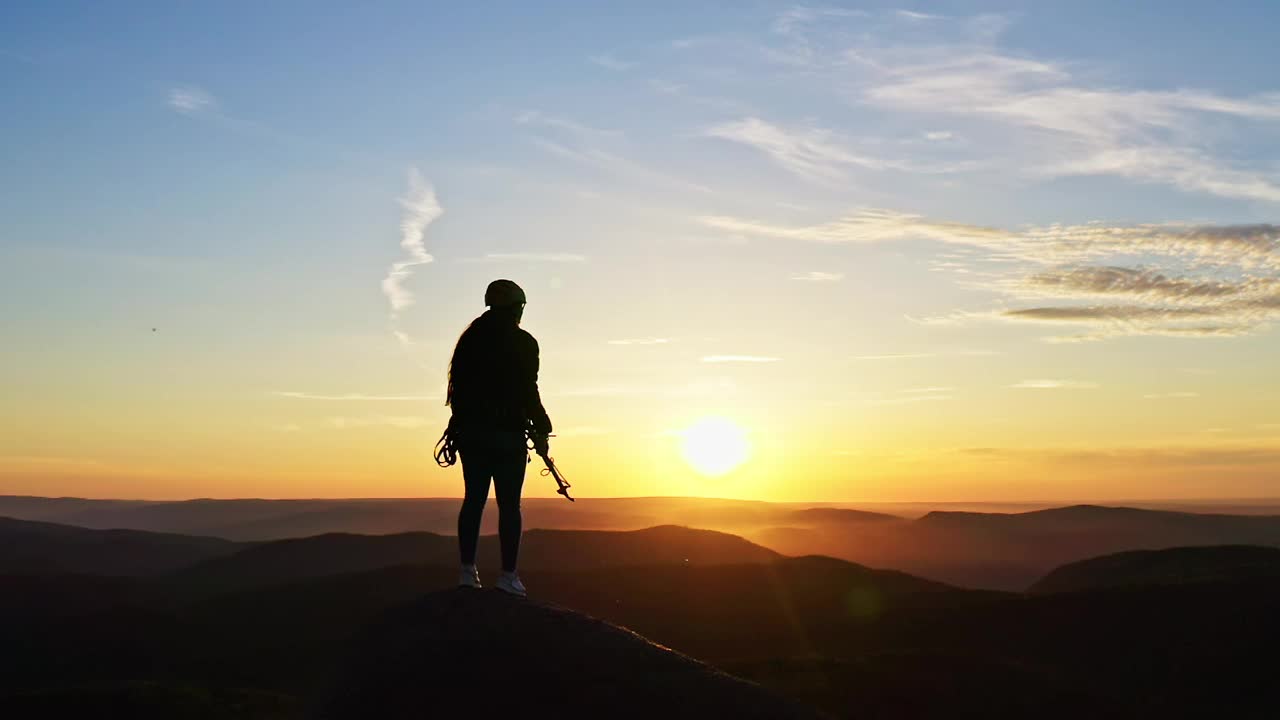 一位年轻的女登山者在夕阳下站在山顶上，胜利地举起双手。视频素材