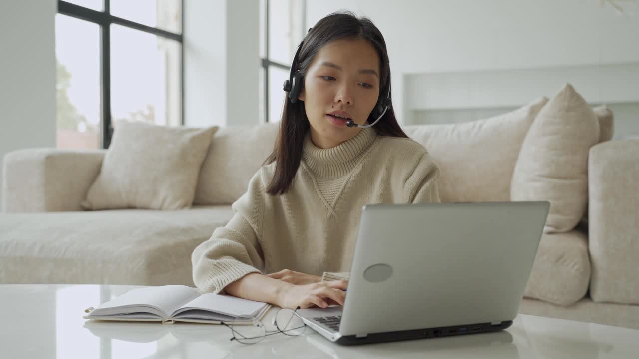 一位亚洲女性戴着耳机看着笔记本电脑屏幕，与对话者交流。韩国女商人戴着耳机视频呼叫客服视频素材