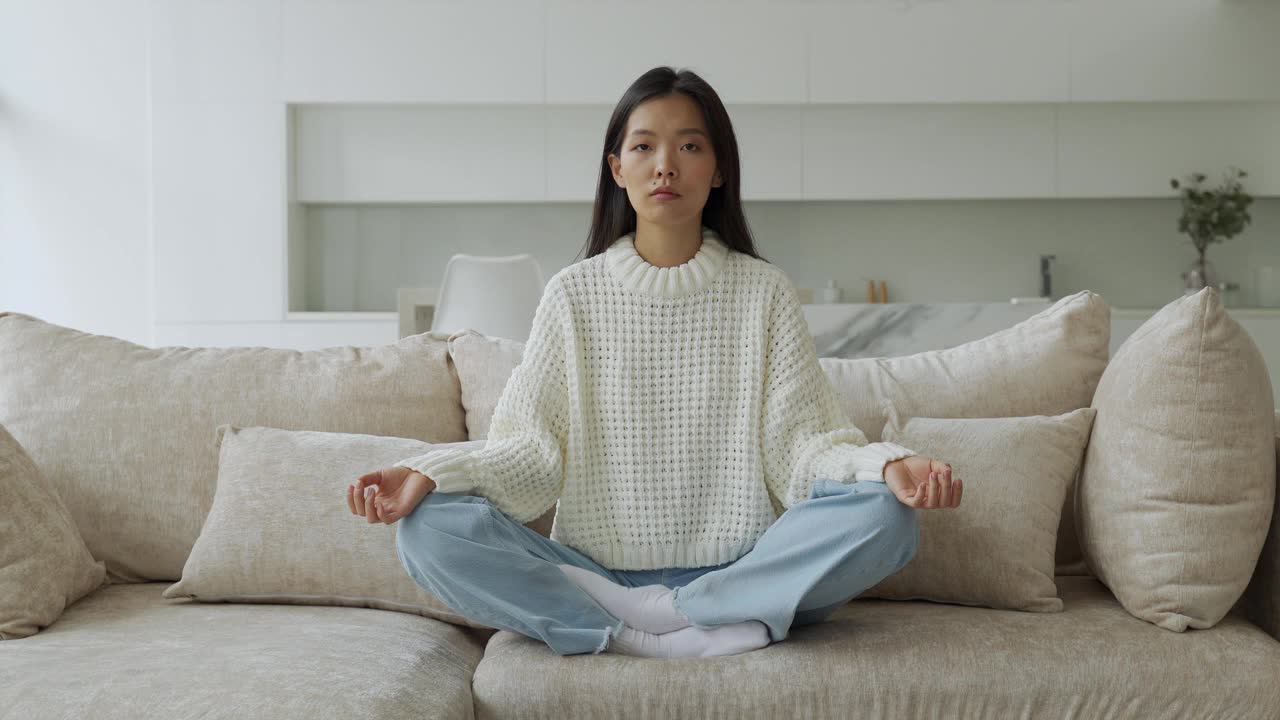 一个亚洲女孩坐在客厅的沙发上做瑜伽。女瑜伽修行者，莲花坐。瑜伽和冥想的概念视频素材