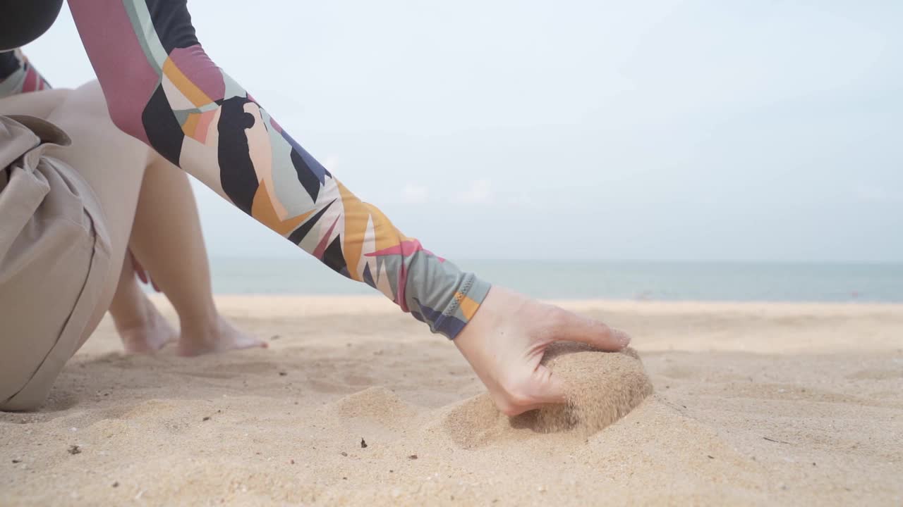 女性在室外沙滩上倒沙子。热带度假插入镜头。慢动作沙粒从游客手中落下。日落晴朗的天空。视频下载