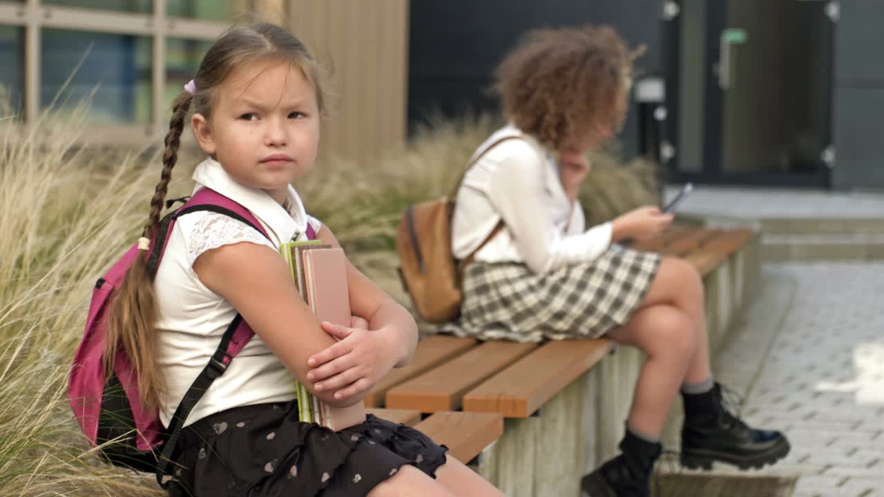 两个年龄不同的女学生坐在学校操场的长凳上。小女孩把书压在胸前，大女孩手里拿着智能手机视频素材