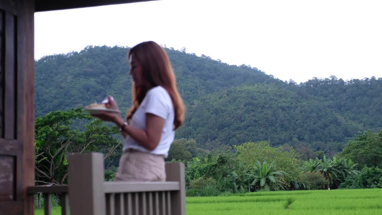 模糊的一个女人吃饭，而站在家里的阳台上，有美丽的稻田和山景视频素材