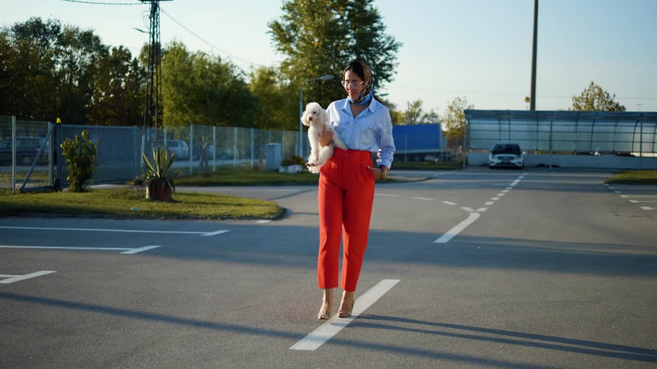 一个漂亮的中年女人抱着一只小马耳他犬走在街上。现代女性带着宠物在城市里散步视频素材