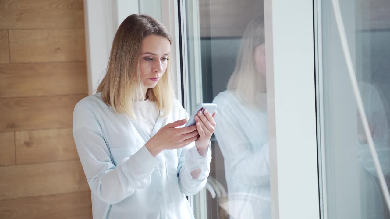 年轻快乐的女子在办公室或家中的窗户边看到了手机上的好消息视频素材