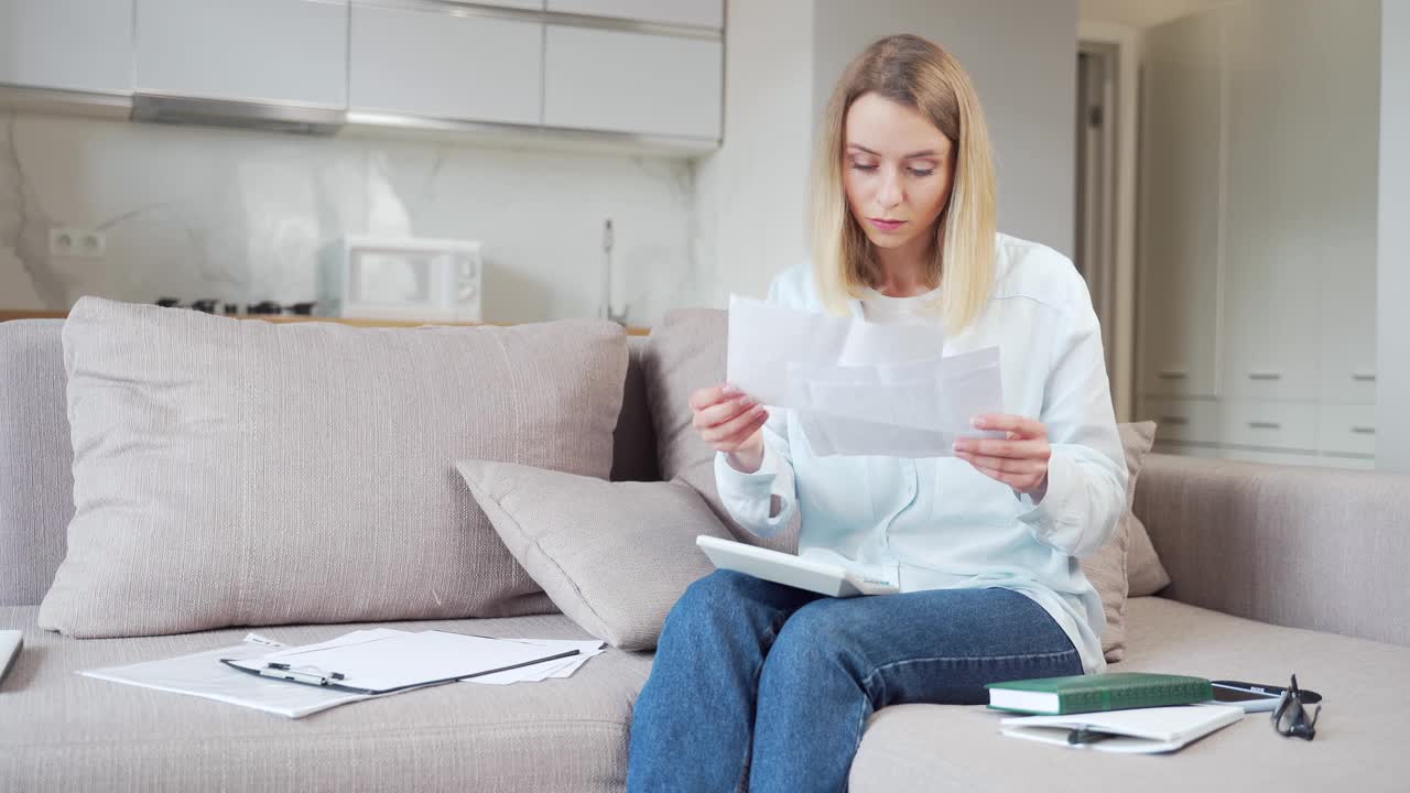 年轻的女性家庭主妇坐在家里的沙发上，处理银行账户、贷款支付和公用事业账单视频素材