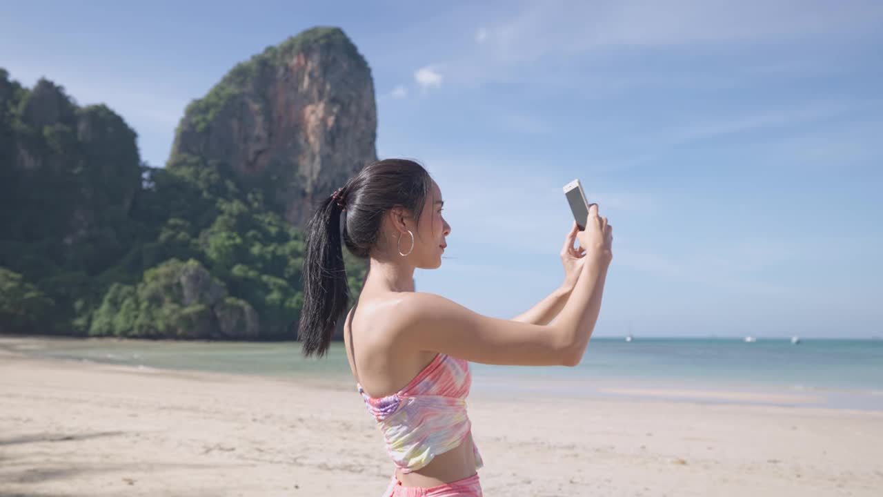 年轻美丽的亚洲女人视频通话与朋友通过智能手机在热带岛屿海滩放松的旅行，无线技术，女士挥手向手机相机显示享受的情感视频素材