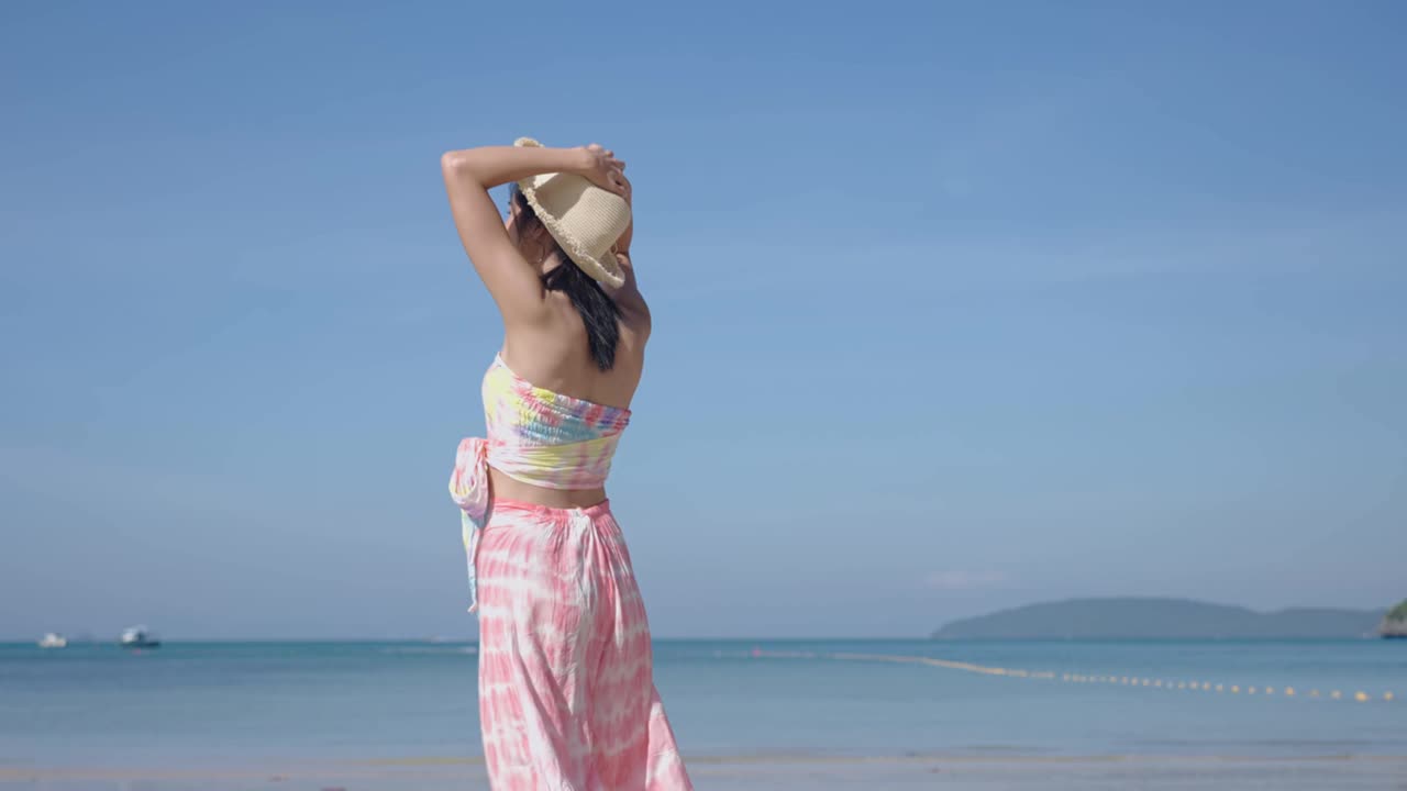 年轻美丽的亚洲女人在夏天的衣服，戴着帽子走在沙滩上与大海的背景。在热带岛屿度假的漂亮女孩。风吹新鲜空气，风吹帽慢动作视频素材