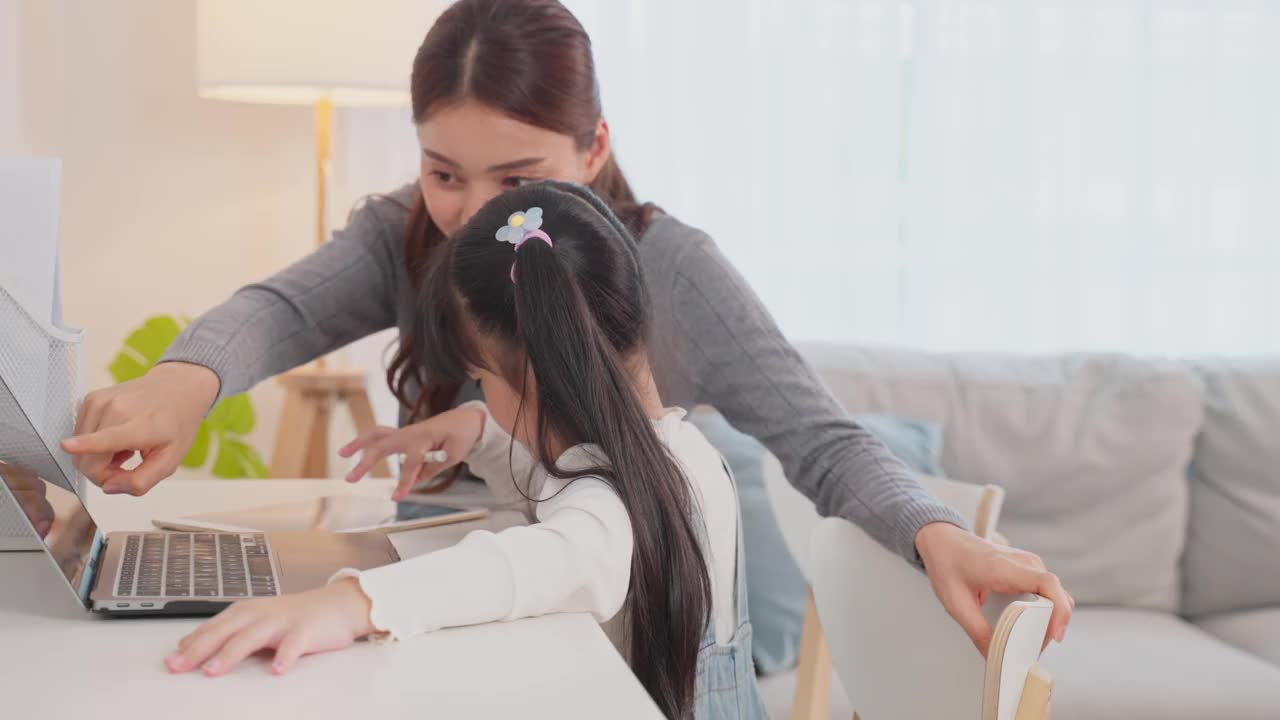 亚洲小女孩和妈妈在家里学习在线课程。学前班的孩子用笔记本电脑做家庭作业，由学校老师通过数字远程互联网在妈妈的支持下完成。视频素材