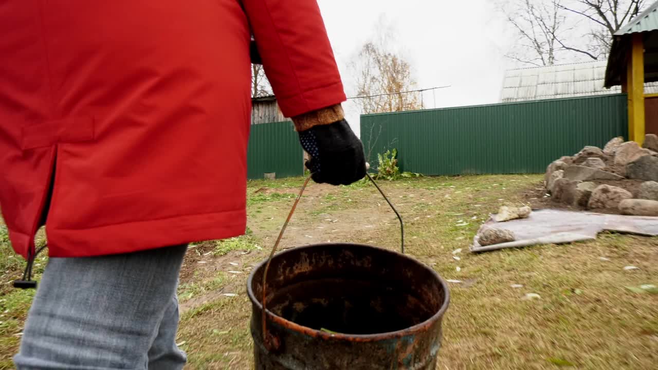 一个穿红夹克的女人从地里移走黄色的落叶。一个女人提着一桶落叶。秋季的家庭视频素材