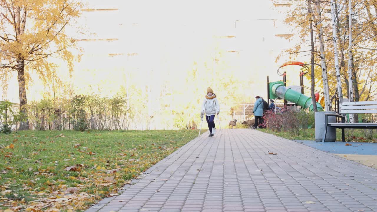 孩子拄着拐杖在秋天公园散步。那女孩打了石膏，一条腿断了。视频素材