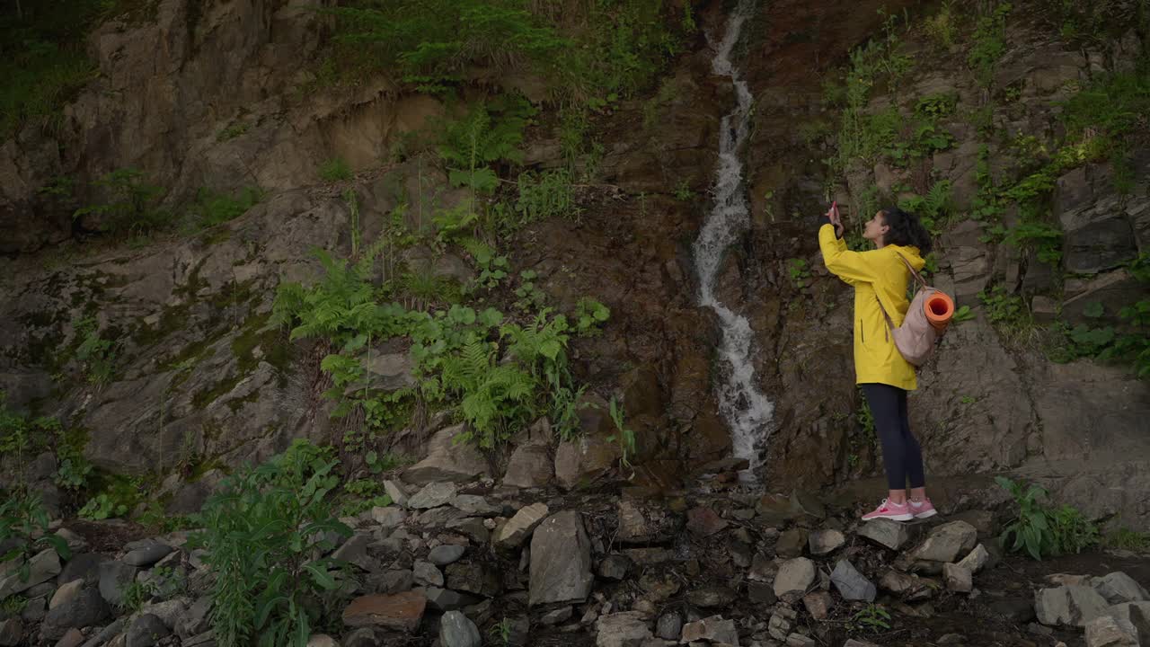 在森林中徒步旅行的女游客正在用手机拍摄瀑布视频素材