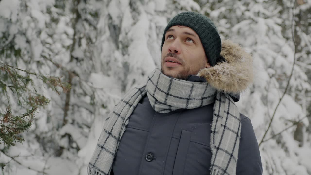 悠闲男子游客的慢动作肖像，站在冬天的森林里，笑着欣赏雪花飘落视频素材