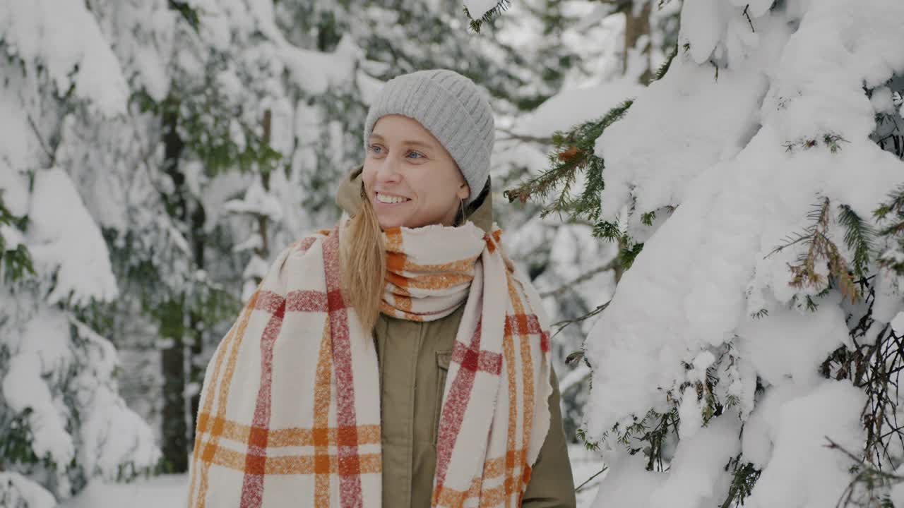 穿着冬衣的年轻女子的肖像，站在松树下的门口，雪花飘落，她笑着视频素材