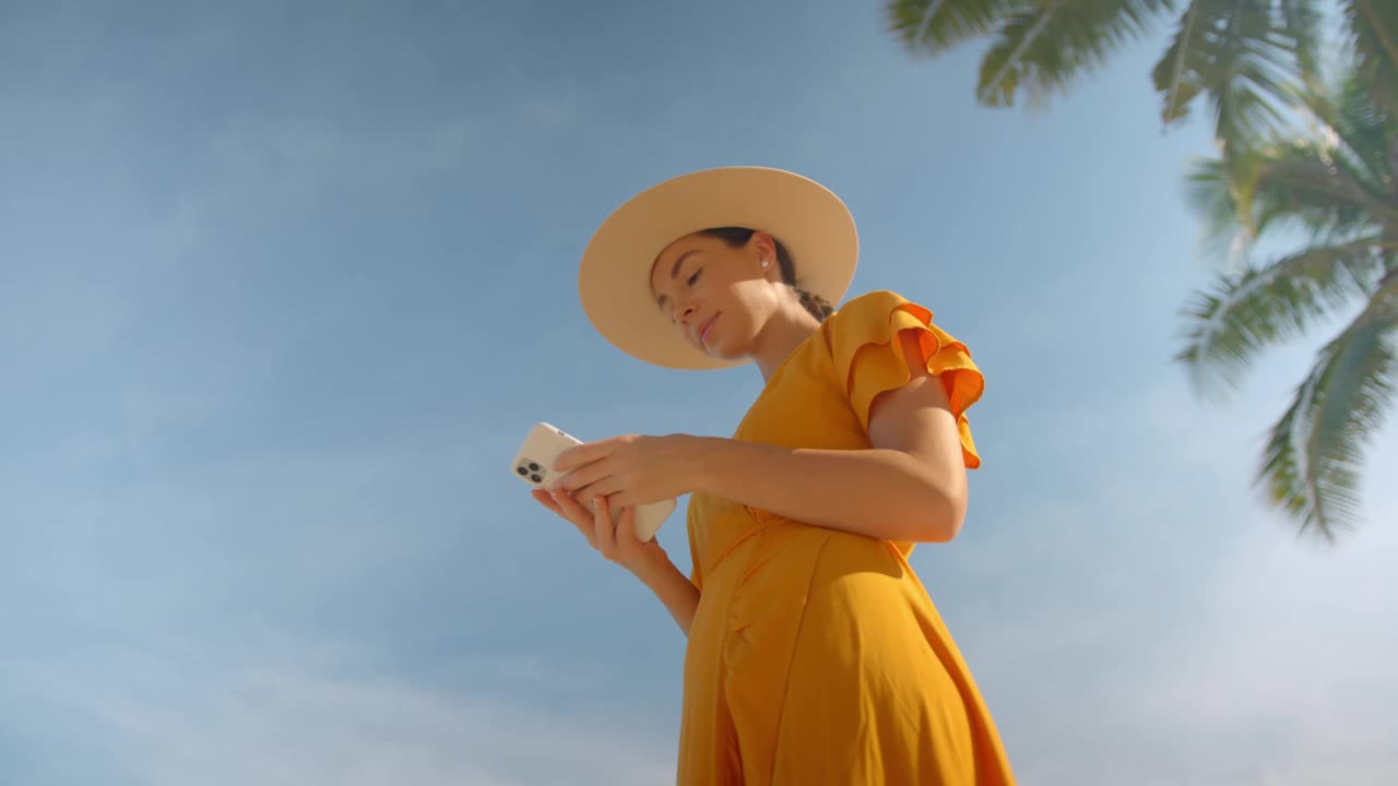 可爱的女人在夏天黄色的衣服和草帽站在棕榈树下，使用她的手机视频素材