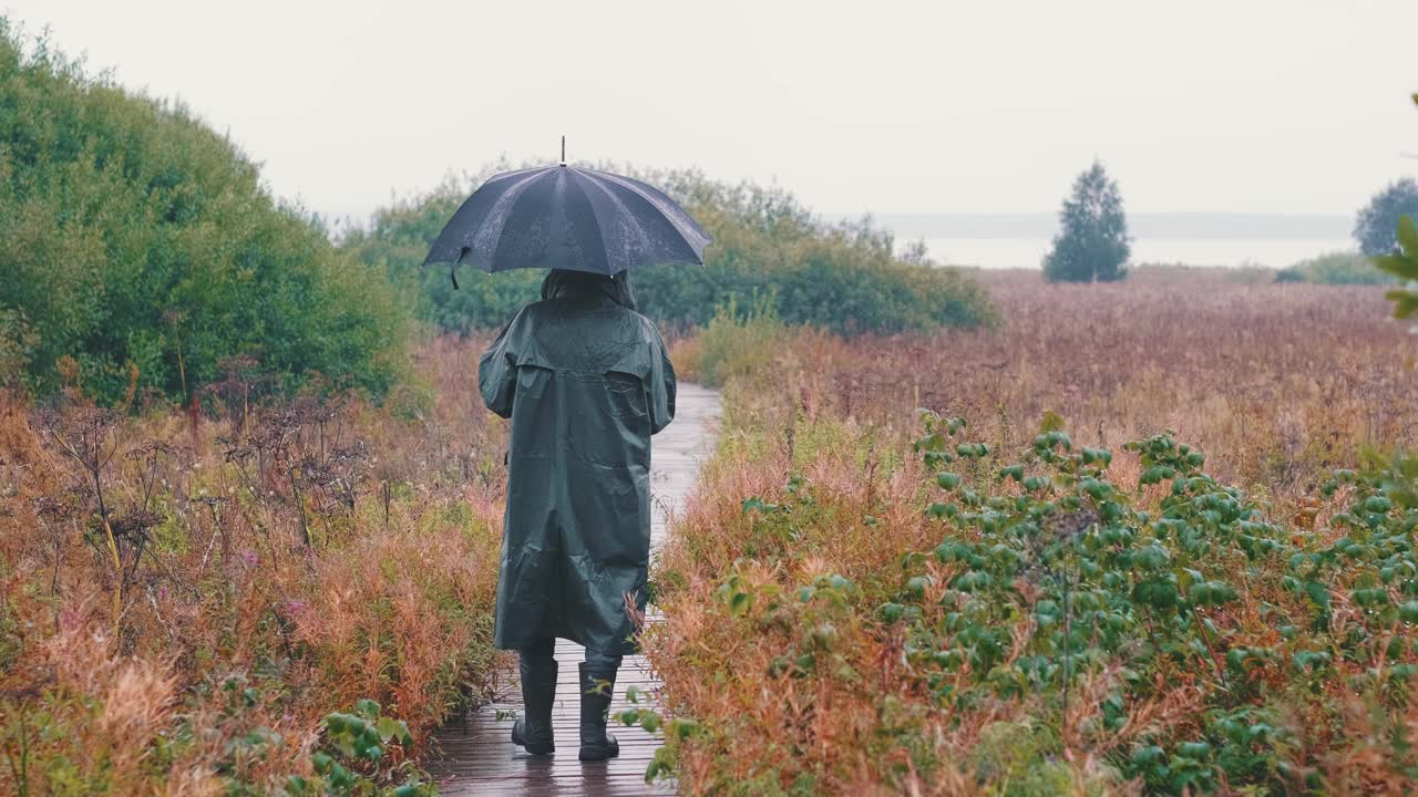 一个悲伤的男人的剪影，穿着雨衣，撑着伞，在雨中走在小路上视频素材