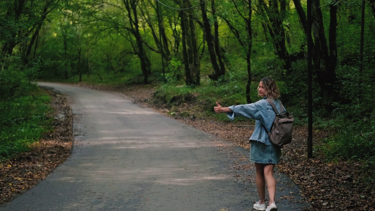 一个美丽的白种人金发女人在休闲的衣服搭便车沿着森林道路。在高速公路边用手抓住一辆车。视频素材