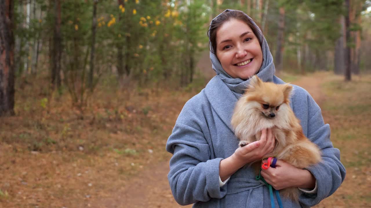 年轻女子抱着波美拉尼亚迷你斯皮兹在秋天公园散步，慢动作视频素材