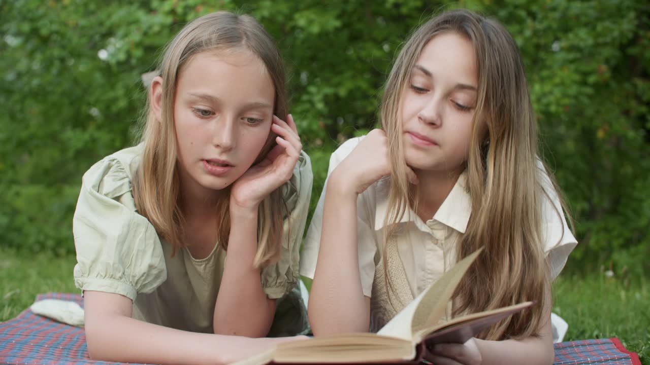 两个年轻的女孩一起躺在夏天公园的绿草地上看书。十几岁的女朋友在暑假放松在绿色的田野上看书页。视频素材