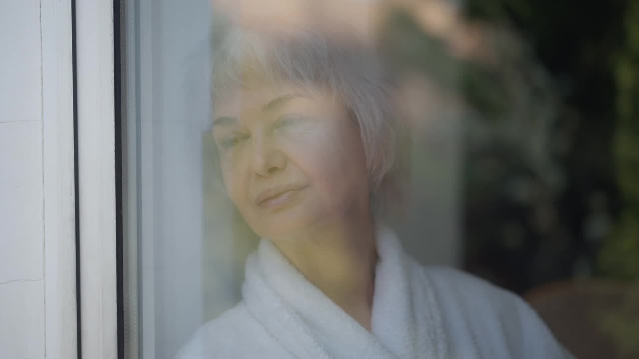 特写成熟的白种人女子看着窗外喝着早晨的咖啡。美丽苗条的女士穿着白色浴衣在家里享受热饮。通过玻璃拍摄。视频素材