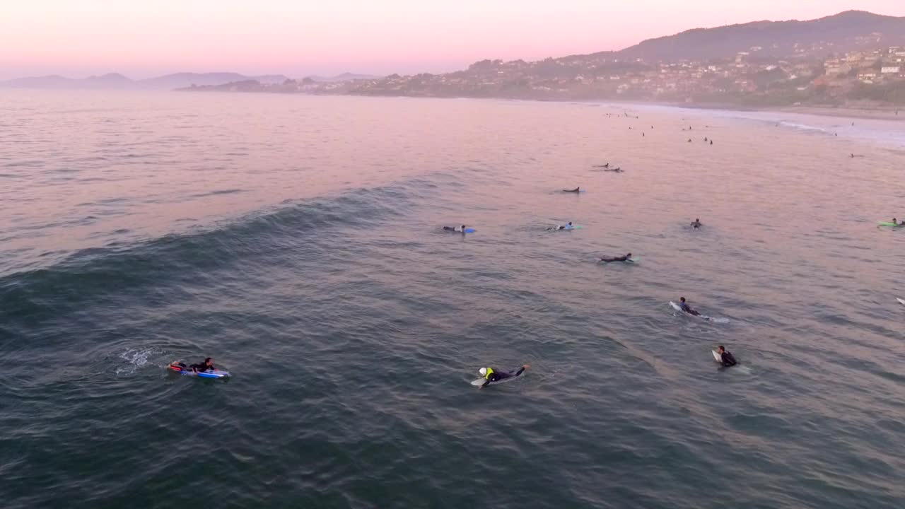 一群冲浪者在板上的海等待波浪骑在粉红色的日落和山坡镇背景洛斯帕托斯海滩，加利西亚，西班牙。空中摄影车左拍。视频素材