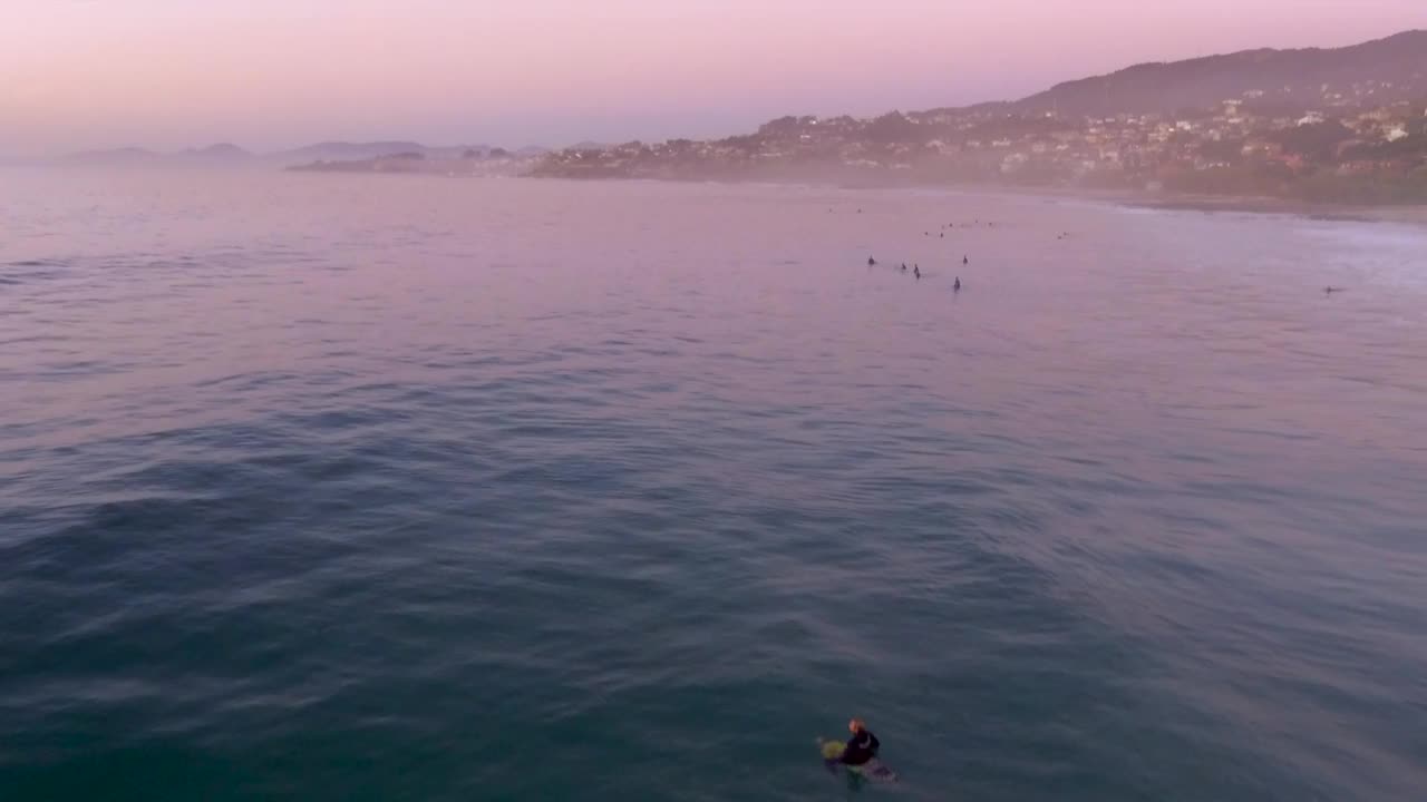 一群冲浪者在板上准备和骑在帕托斯海滩上粉红色的日落和山坡镇背景，加利西亚，西班牙。空中拔射。视频下载