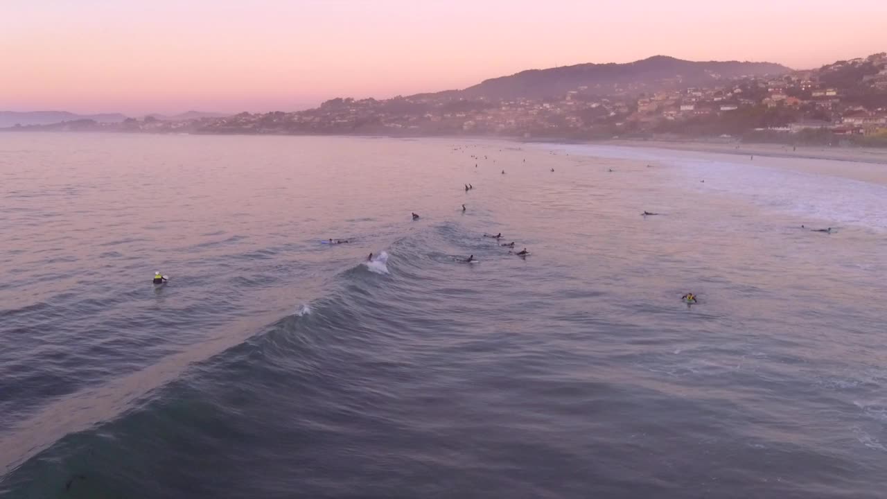 一群冲浪者在帕托斯海滩上粉红色的日落和山腰镇的背景，加利西亚，西班牙。天线跟踪拍摄。视频素材