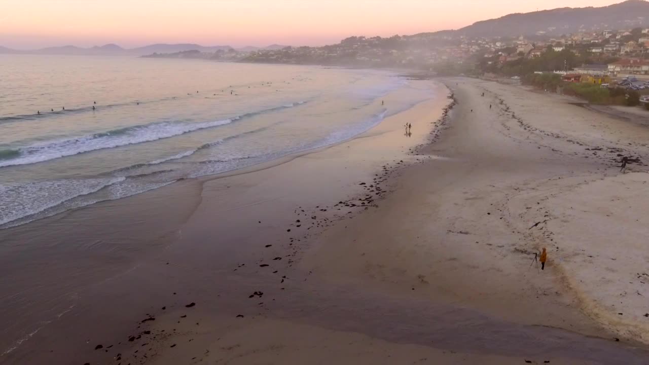 鸟瞰图的波打破在帕托斯海滩和冲浪者在海上等待骑在金色的日落和山腰城镇背景，加利西亚，西班牙。退出。视频素材