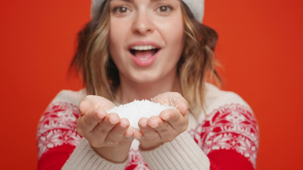 快乐的年轻女子从她的手吹雪到相机在圣诞服装圣诞帽特写慢动作在红色的背景。冬天的节日气氛。积极的情感。的生活方式。圣诞节的气氛视频素材