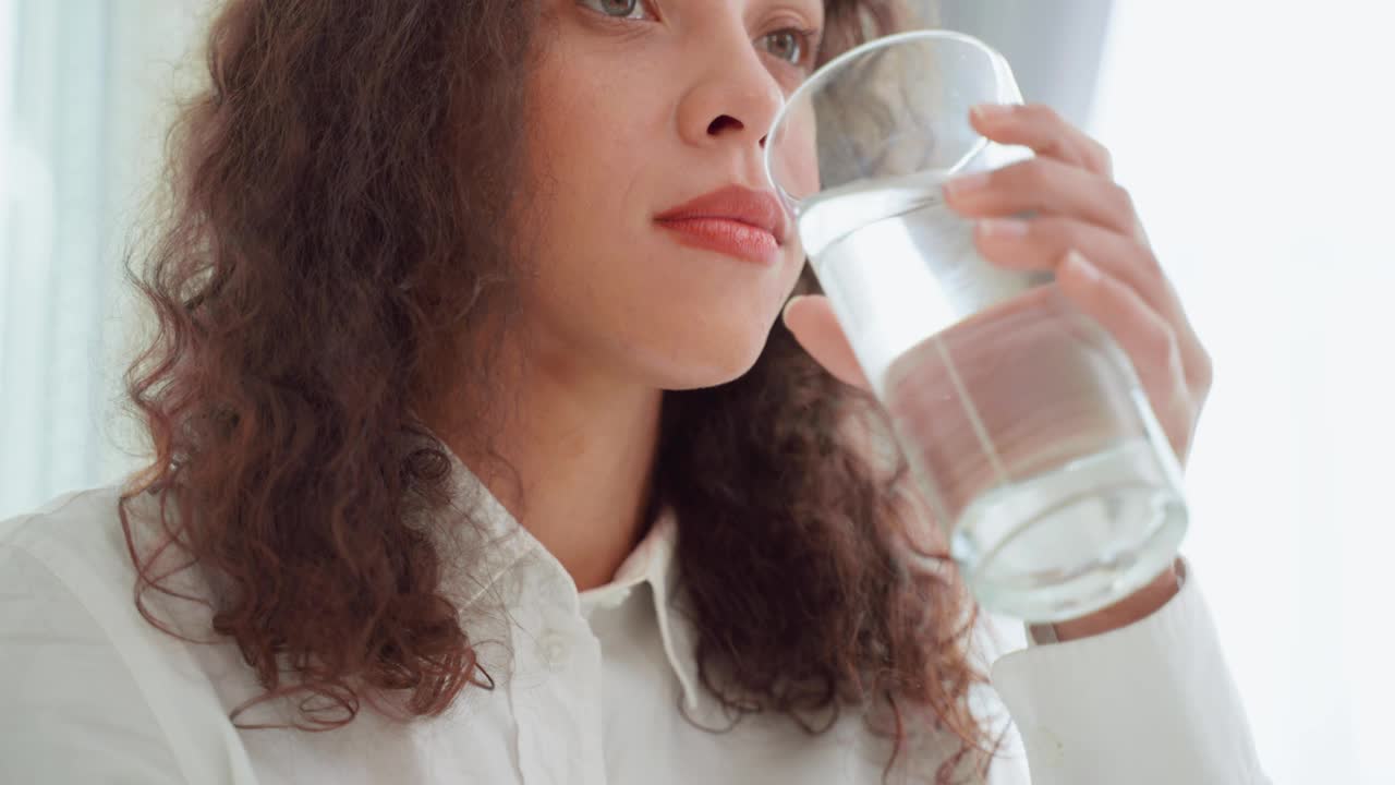 拉丁美洲女人的肖像坐在床上，拿着一杯水在卧室。年轻干渴迷人美丽的女孩早晨醒来后在家中为健康啜饮一杯洁净的矿物质天然视频素材