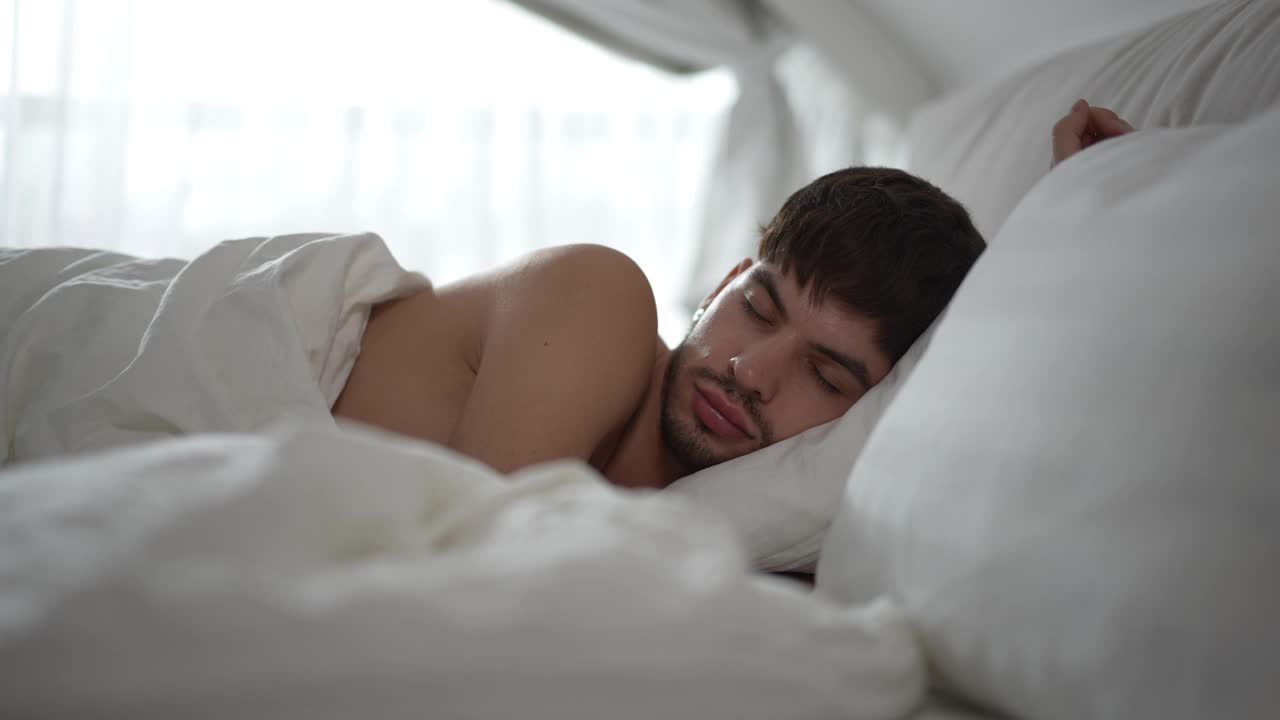 熟睡的年轻白人英俊男子在舒适的白色床上转动着眼睛。大胡子早上在家里舒适的卧室里醒来。缓慢的运动。视频素材