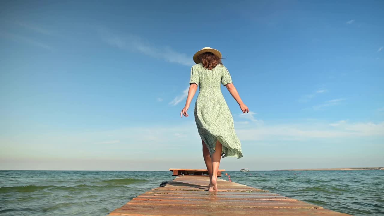 在海风吹拂的清晨，一位身穿绿色夏装、头戴草帽的年轻女子慢动作地沿着木栈桥走着。走路优雅、美丽、柔美视频素材