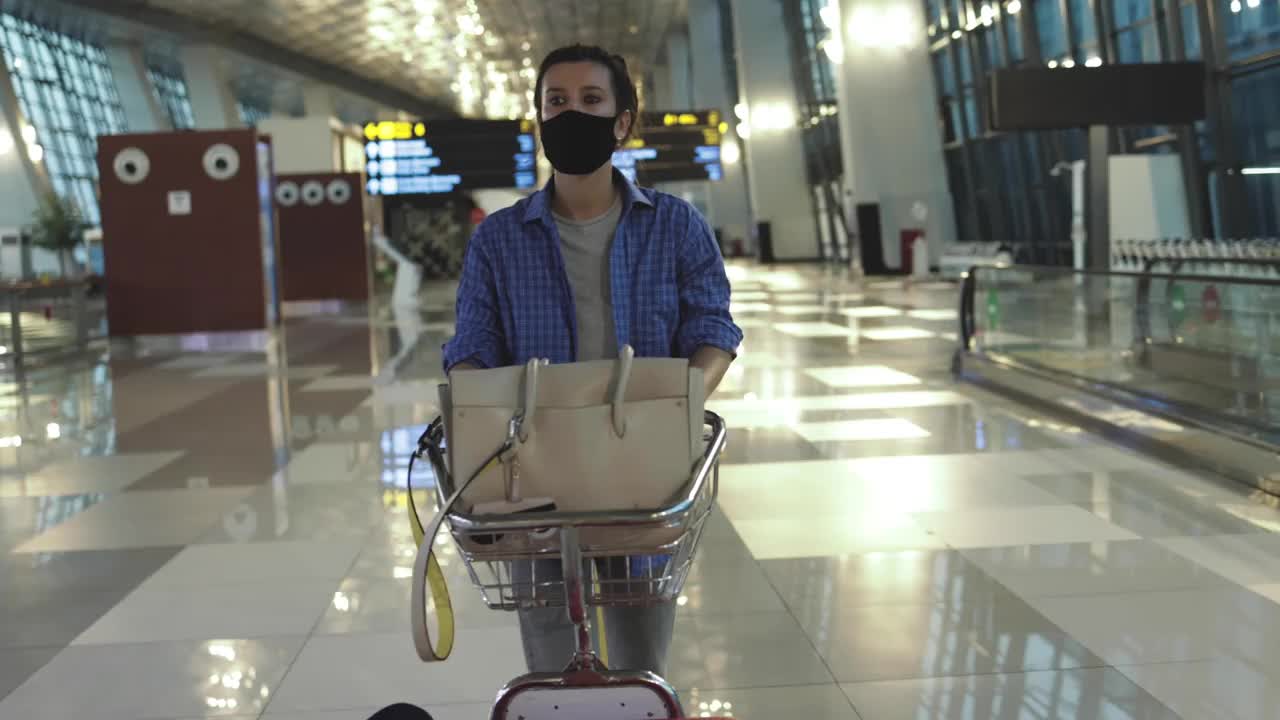 戴着口罩的年轻女子在机场候机楼推着行李车走着。流行病和旅行的概念视频下载