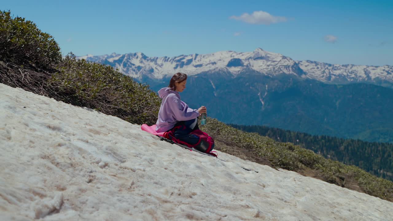 登山途中，一位年轻女子在山坡上停下脚步，一边喝水一边欣赏风景视频素材