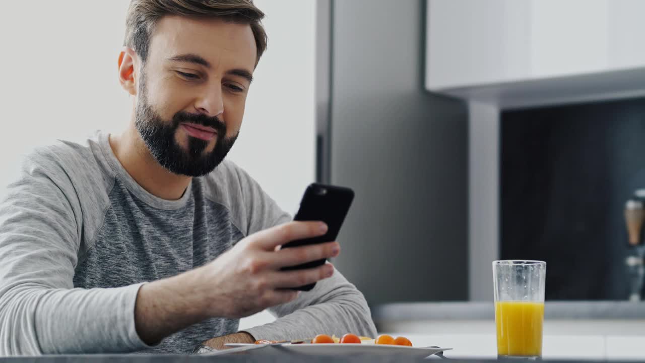 近景:微笑的大胡子男人在吃晚饭和使用智能手机视频素材