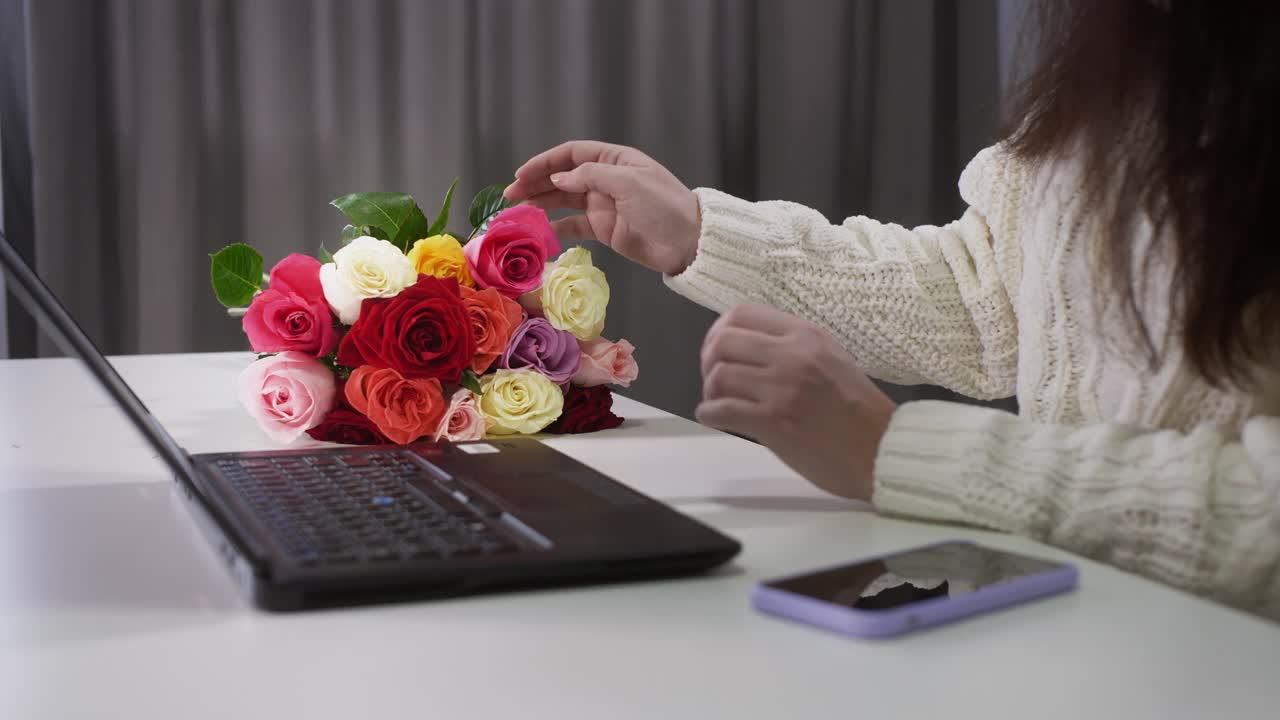 一位女士在工作场所触摸玫瑰花束。一名女子坐在办公桌前，手里拿着笔记本电脑、智能手机和美丽的新鲜玫瑰视频素材