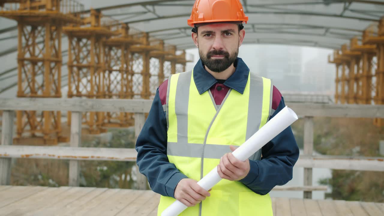 男建筑工人像，头戴钢盔，身穿背心，手持图纸站在工地外视频素材