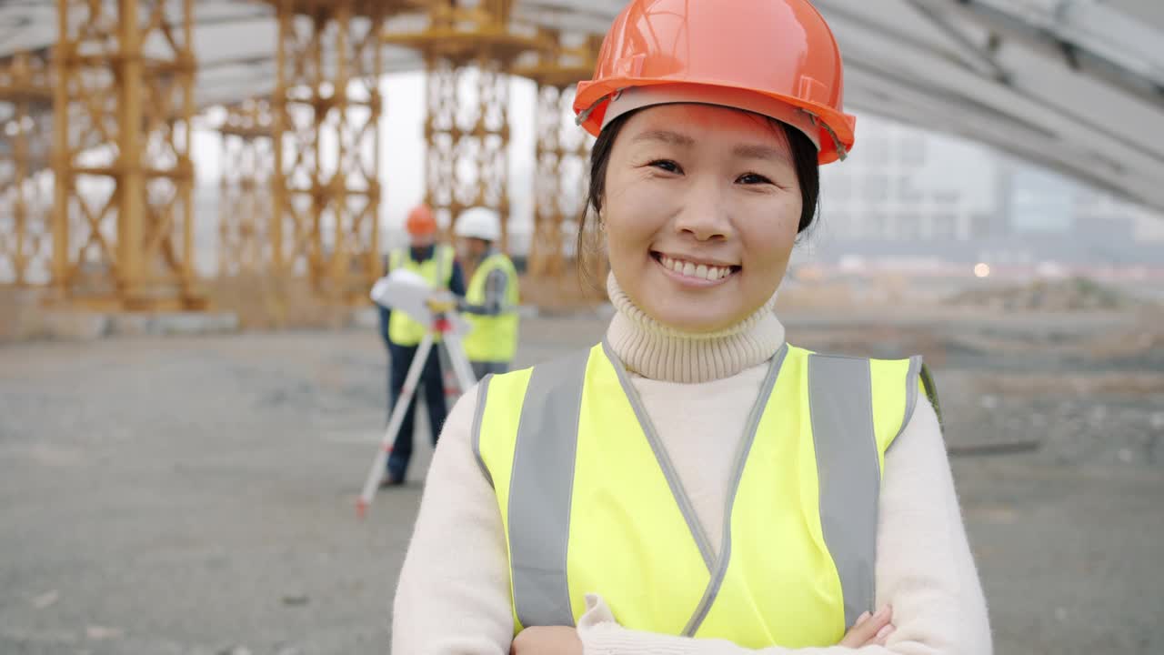 身穿制服的亚洲建筑工人笑容满面地站在工地上，人们在一旁工作视频素材