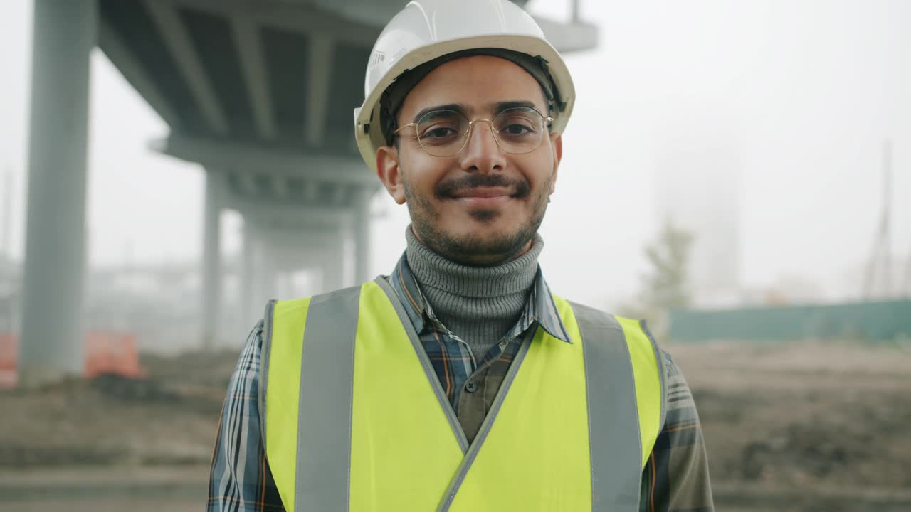 阿拉伯男子的肖像，戴着头盔和背心微笑站在城市建设的背景视频素材