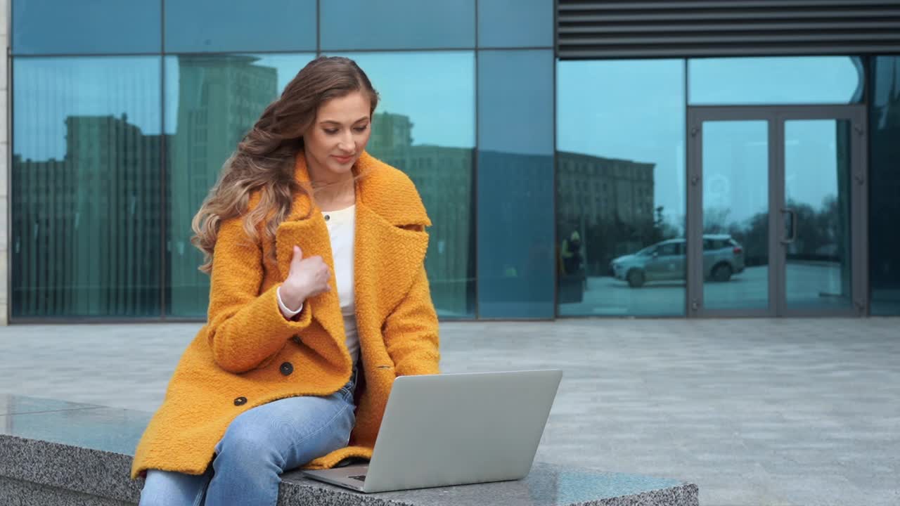 商务女性穿着黄色外套，穿着笔记本电脑坐在公司大楼的室外背景视频素材