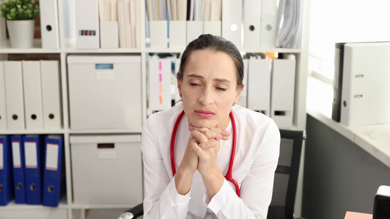 脖子上戴着听诊器的悲伤女医生坐在诊所的桌子旁看4k电影视频素材