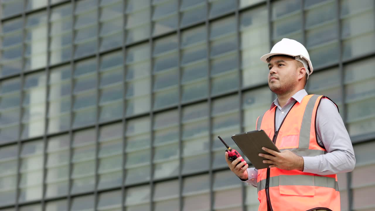 亚洲工程师手持无线电指挥工人施工。年轻的印度建筑工人在现代建筑工地用收音机和平板电脑交谈视频素材