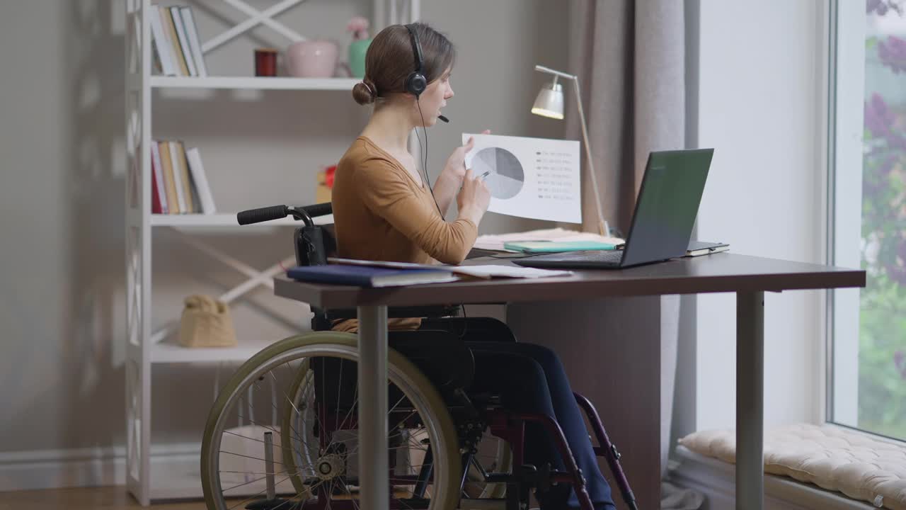 自信的白人年轻女子的肖像在轮椅上，在笔记本电脑视频聊天指着图表。美丽苗条的自由职业者工作消息在线从家庭办公室。远程工作的自由。视频素材