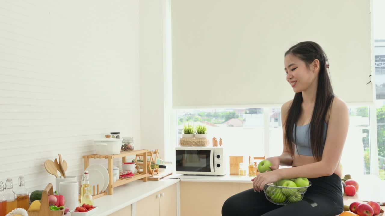 快乐的亚洲健康女人在公寓的厨房里拿着篮子吃着青苹果。视频素材