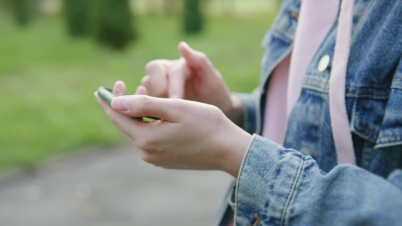 一个穿着牛仔夹克和粉色连帽衫的女人的手的特写在公园里使用智能手机视频素材