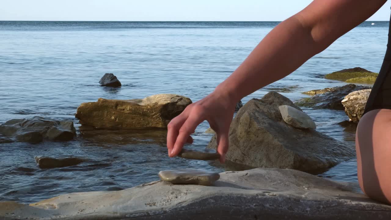 穿着黑色泳衣的年轻女子在岩石海岸堆石头。女人平衡岩石在大海或大海在日出。瑜伽和平衡的概念视频素材