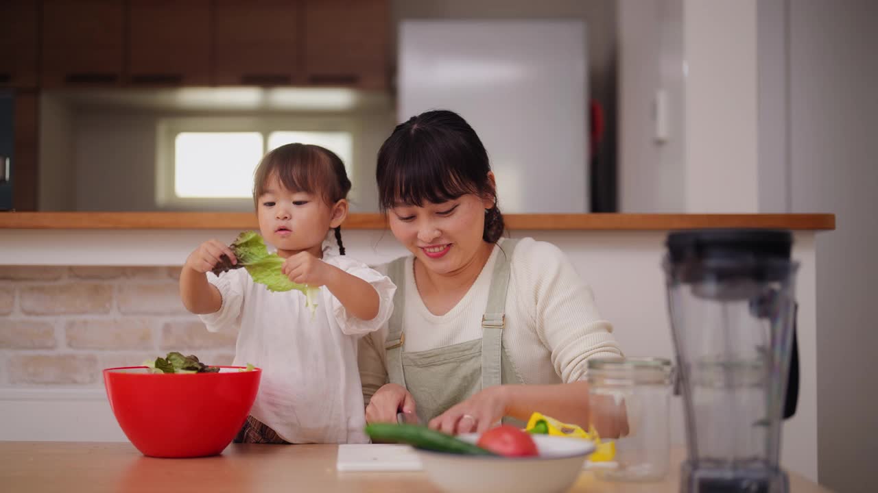 母亲和她的小女儿在家里准备沙拉。妈妈帮她的小女儿准备蔬菜，鼓励她健康饮食视频素材