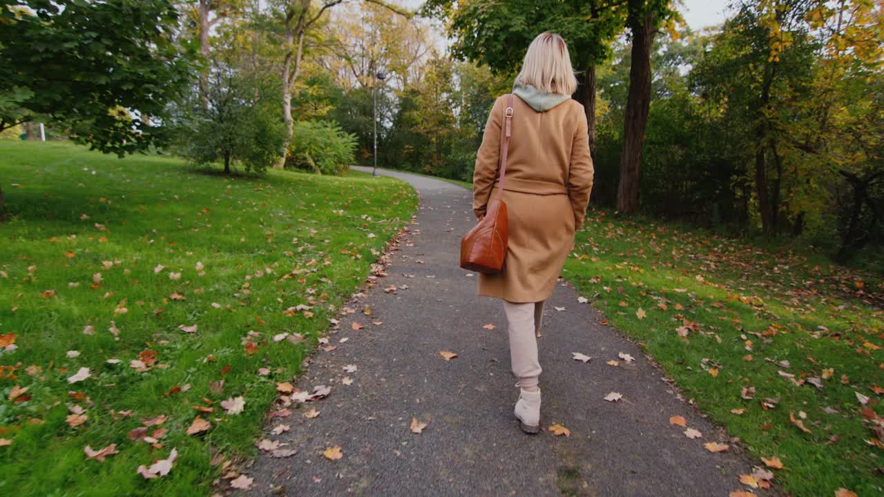 一个女人穿着外套，拿着一个袋子在秋天的公园里散步，后视图视频素材